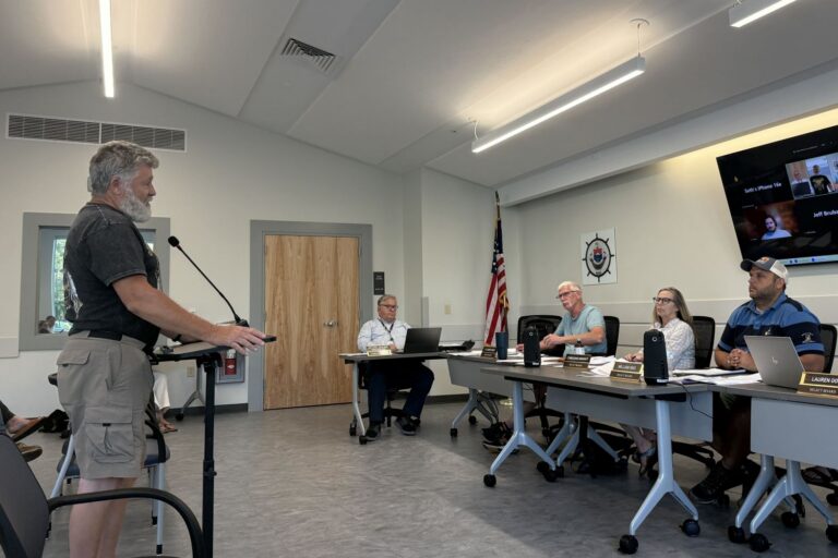 a man stands at a podium while speaking to the select board.