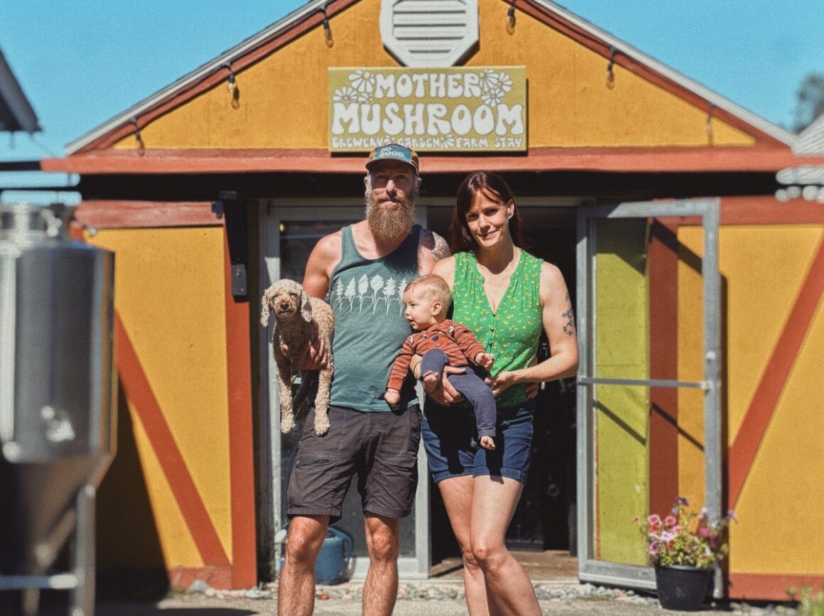 a man and woman standing in front of the entrance to a brewery. the woman is holding a baby in her arms while the man is holding a dog.