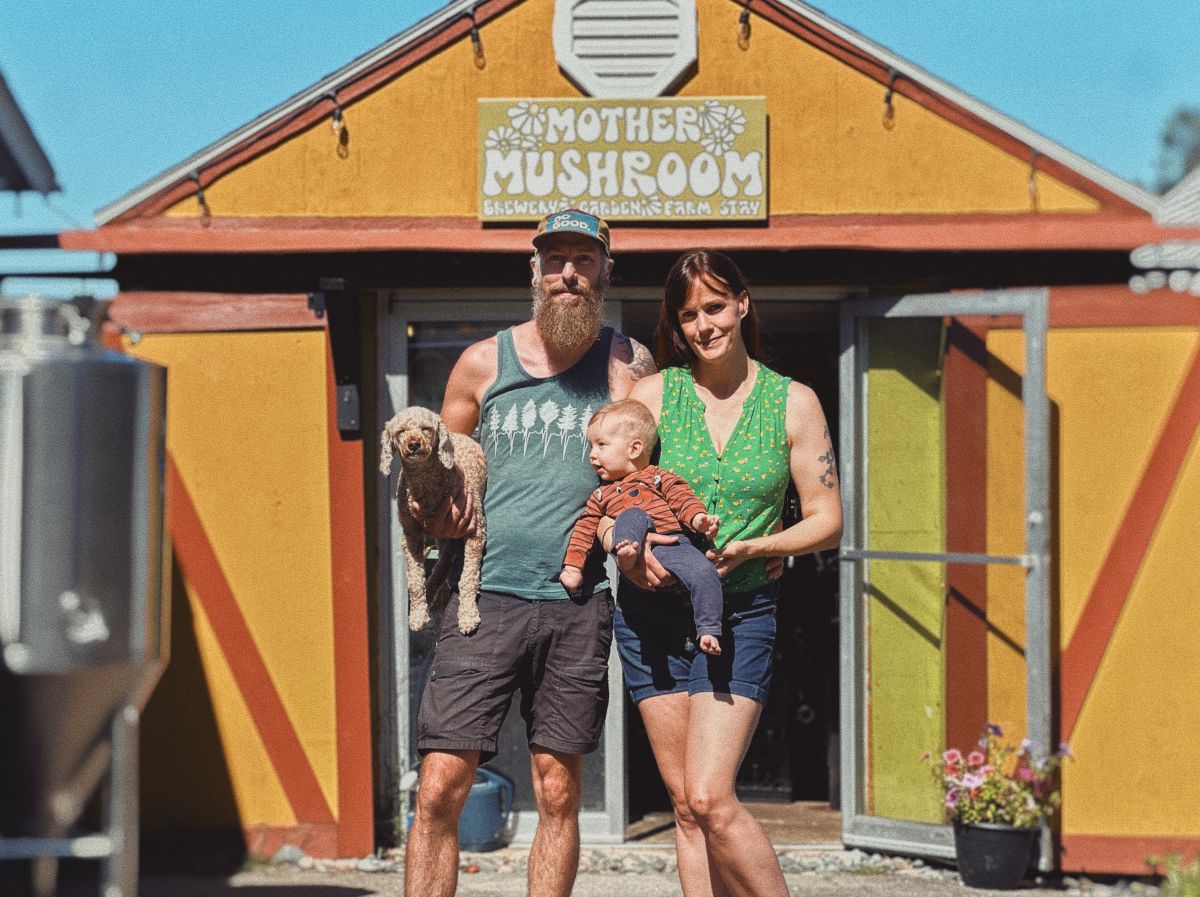 a man and woman standing in front of the entrance to a brewery. the woman is holding a baby in her arms while the man is holding a dog.