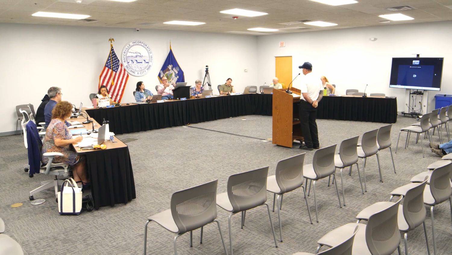 members of the city council sit at a u-shaped table while listening to a speaker standing at a podium
